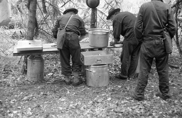 Three soldiers stand with their backs to the camera.  Two of them are leaning over a table that has boxes and pots of food on and beside it.  Trees are seen in the background.