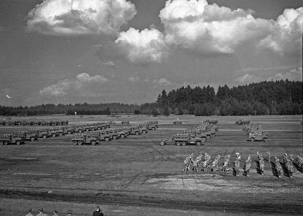 An elevated landscape view of military trucks and soldiers in lines in a wooded clearing.