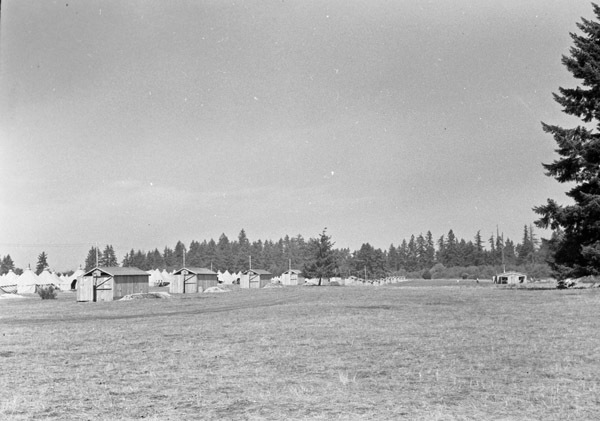 A semi-permanent military camp along a dirt road in a wooded clearing.  Some small wooden buildings appear closest, with rows of large tents behind them. 