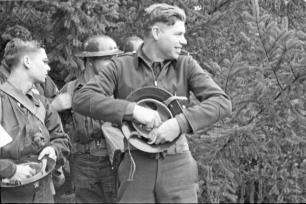 A soldier smiling and looking to the right of the camera while holding and possibly adjusting their helmet.  A few other soldiers are directly behind him with trees in the background.