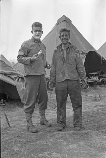 A pair of soldiers standing and looking at the camera humorously. The man on the left is holding shaving equipment and has shaving cream covering the bottom of his face. The man on the right appears to be disheveled and dirty, while smiling.