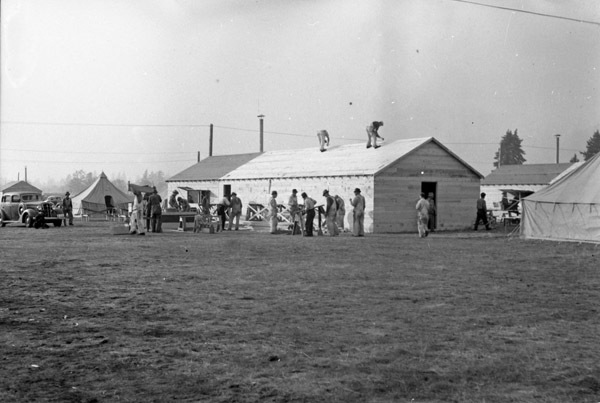 A couple dozen men working on a building that is nearly complete. Two men stand on the roof, while the rest work on the ground.