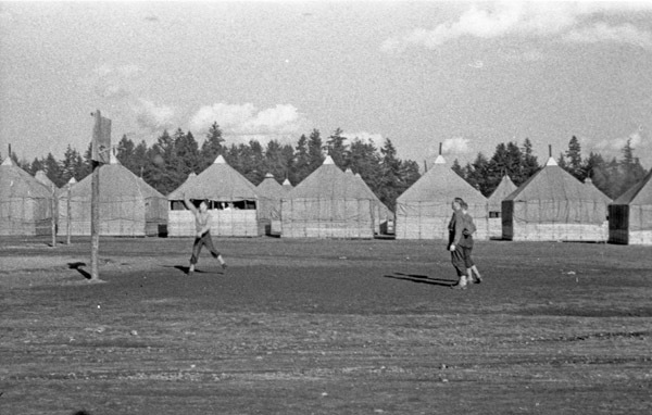 Three soldiers playing basketball in a field.  Tents or small buildings line the background, with trees behind the building.