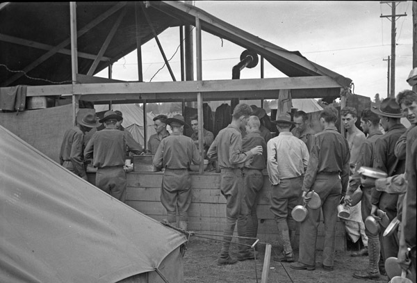 A line of soldiers in front of a temporary wooden building being served food by those within the building.
