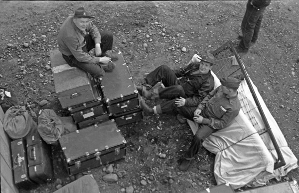 Three soldiers lounging on storage trunks and bedding that is stacked outdoors.