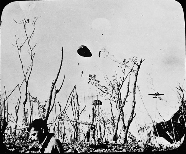 A couple of paratroopers can be seen in the sky with their parachutes fully deployed. A military plane is also in the sky near them. In the foreground there are a few soldiers standing by some desolate foliage.