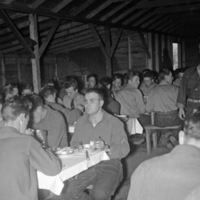Dozens of soldiers sitting at eating at long tables in a mess hall.