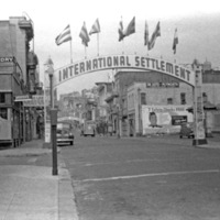 A city street with an arch that reads, "International Settlement" with numerous flags flying atop it.  The street has a few cars parked along it.