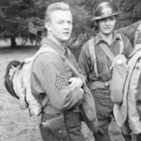 Two soldiers standing and looking at the camera.  The man on the left has their helmet off, while the one on the right is wearing their helmet.