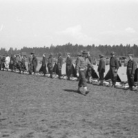 A line of soldiers standing with their backs to a line of tents, apparently preparing for inspection. In front of the tents are neatly organized groups of equipment, most likely the contents of the tent and the soldiers' belongings.  A single man is walking along the line of standing soldiers.