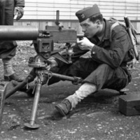 A soldier sitting on a seat behind a tripod-mounted machine gun.  He is looking through the sights of the gun.