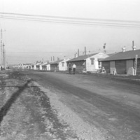 A line of single story houses on the right, with a dirt road on the left.  Two people can be seen walking down the road with their backs to the camera.
