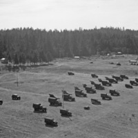 More than a dozen military trucks lined up in 3 lines, some of which have artillery cannons hitched to the back.  A tent camp and a forest are in the background.