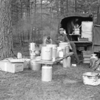 Three soldiers are organizing pots and boxes of food on a table and taking them off or on a military truck.  They are in a heavily wooded area.