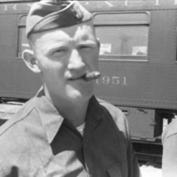 A soldier wearing a garrison cap and smoking a cigar facing the camera.  A train car with "1951" printed on it can be seen in the background.