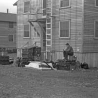 A soldier sitting on a stack of storage trunks outside of a barracks.  Other stacks of trucks are also seen.