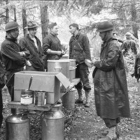 Four soldiers stand on the left of a table with pots and boxes of food.  Two men stand to the right, the closest wearing a rain poncho.