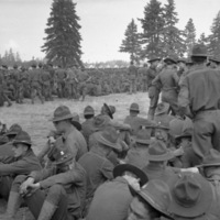 Hundreds of soldiers sitting or standing in relaxed positions in a field.  Some fir tress are seen in the background.