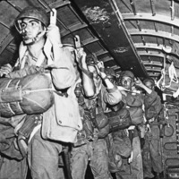 A half dozen soldiers in full paratrooper gear standing in the interior of an airplane holding to hooks that secures them in place.  They look to be getting ready to parachute out of the plane.
