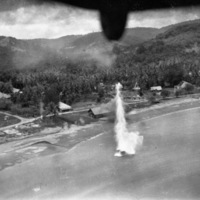 A plume of water showing a bomb exploding in the water near a shoreline populated by houses and other buildings.