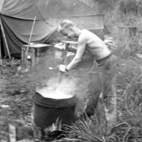 A shirtless soldier uses a pole to to stir clothes in a steaming bucket.  A tent and trees are in the background.