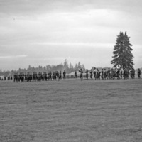 A line of soldiers playing musical instruments in a parade formation marching.  More soldiers and tents can be seen in the background.