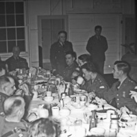 A group of soldiers sitting at a long table having a dinner.  While some are standing, most are sitting while eating and drinking.