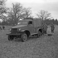 A military truck in a wooded clearing, facing just the the left of the camera.  A few soldiers are standing near the back of the truck, while a few others are sitting down near a tree to the left of the truck. 