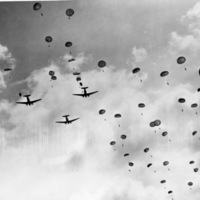 Three military planes in the sky flying away as seen from below. There are numerous paratroopers in the sky behind the planes in various stages of deploying their parachutes.