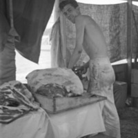 A shirtless man in a tent standing and looking at the camera over his shoulder while cutting a large slab of beef that is on the table beside him.