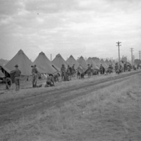 A long row of large tents along a dirt road.  Dozens of soldiers are standing or kneeling outside the tents, looking to be working.