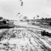 A soldier laying in the mud being pulled by his parachute.  Many more paratroopers can be seen still landing in the background.