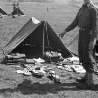 A soldier stands in front of a tent, where a large number of items, presumably the soldier's equipment,  are arranged in front of the tent.  Tents can be seen on either side of this one, as well as behind it, with similar equipment organized in front of them.