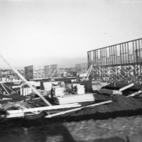 Multiple buildings in the very early stages of construction.  Only one wall per building has been raised, and large wood piles are around each of them.