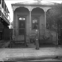 A soldier in dress uniform and a camera hanging from their neck standing in front of a single-level house.