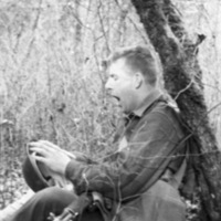 A soldier sitting on the ground with their back against a tree.  They look to be yawning. and holding their helmet in their lap.