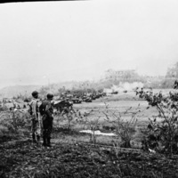 Two military officers look over a field of artillery.  Smoke and destroyed buildings are in the background, likely from a recent battle.