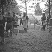 A group of soldiers in a wooded clearing standing around pots of food.  The man in the center is wearing a white cook's apron and hat.