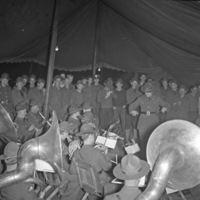 A military band plays inside a large tent.  One man is conducting, and the entire background is filled with more soldiers watching the performance.