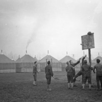Eight soldiers playing basketball around a hoop.  The ball can be seen in mid-air hitting the backboard.  Small houses or tents line the background.