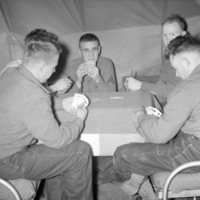 A group of five soldiers sitting on a pair of cots and playing card games on a card table that is set up between them.