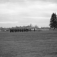 A distant view of a dozens or more soldiers in formation while officers walk in front of them. A tent camp and milling soldiers can be seen in the background.