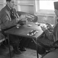Two soldiers sitting on bunks with a card table between them, playing cards and smoking.