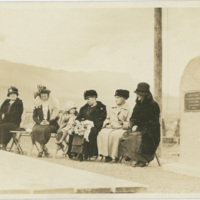 A group of six women and one young girl sitting on chairs next to a dedication plaque.