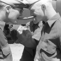 Two soldiers humorously face each other while faux-scowling.  Each of them has a cigar in their mouths, and the cigar ends are touching each other.  Another soldier is behind them and peering at the seen amusedly.