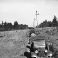 A line of military trucks on a dirt road which runs adjacent to electrical poles.  Recently cleared trees can be seen on the right, along with uncut trees in the background.  