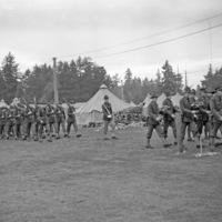 Two large groups of soldiers marching in formation next to a tent camp.  The far right group is in front and holding musical instruments.  The leftmost group in the back has rifles resting on their shoulders.  