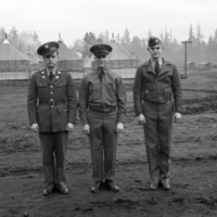 Three soldiers in military dress uniform standing shoulder-to-shoulder posing for the camera.  Tents and fir trees can be seen in the background.