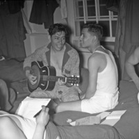 A group of five Hispanic soldiers sitting or laying on the floor in casual clothes and smiling.  The soldier in the middle is playing a guitar. 