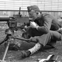 A soldier sitting on a seat behind a tripod-mounted machine gun.  He is looking through the sights of the gun.
